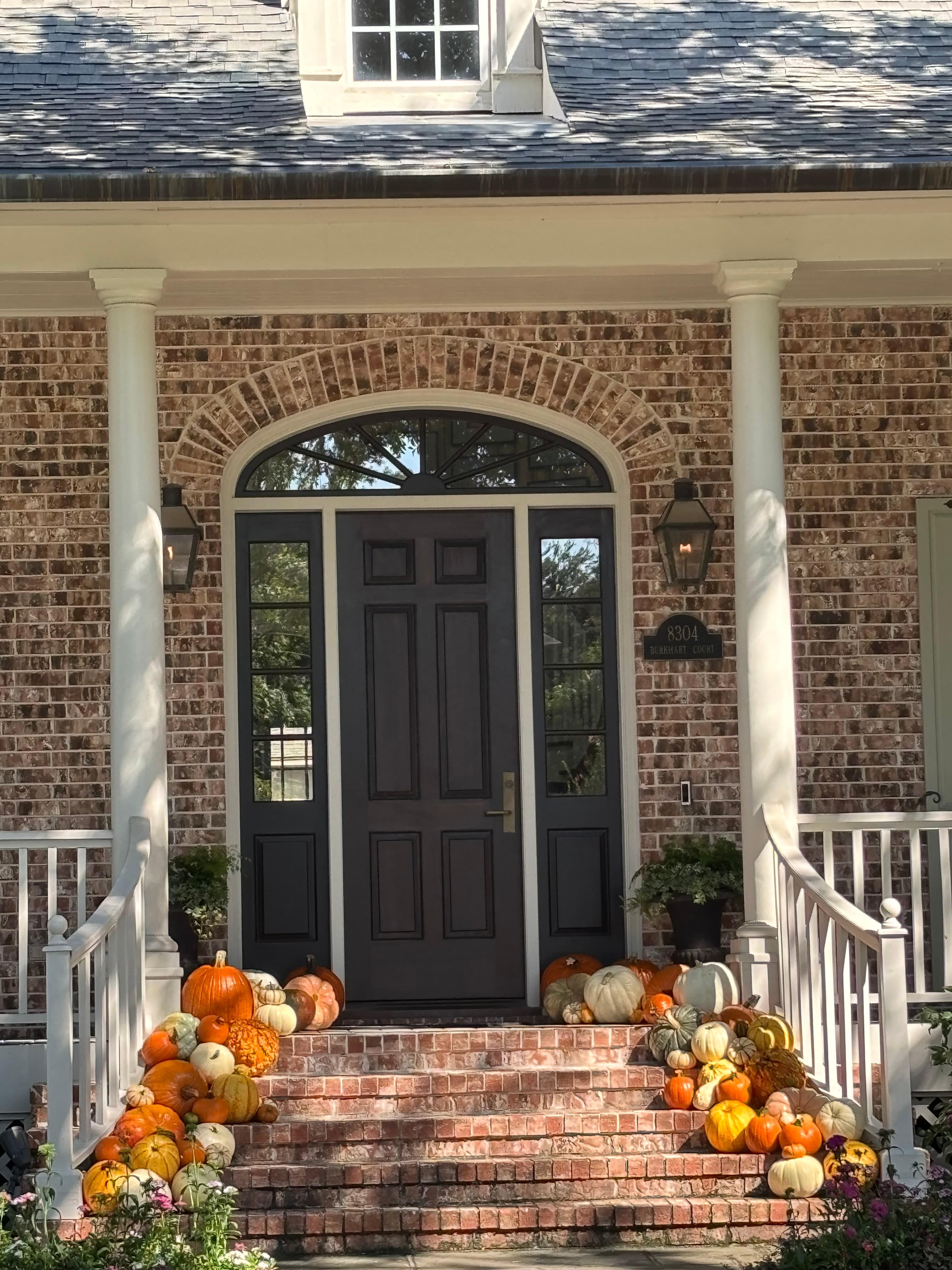 Various pumpkins and gourds decorate the brick steps leading to a dark arched front door.